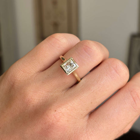 Gold Vintage-inspired, princess-cut diamond rub-over solitaire engagement ring on a person's finger against a neutral background