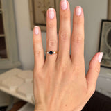 Hand wearing a Vintage, 1960s mid-night blue sapphire & diamond three-stone engagement ring in yellow gold against a blurred indoor background
