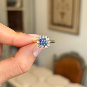 A hand holding an antique-inspired gold ring with a central blue sapphire and a halo of diamonds, set against a softly blurred indoor background.