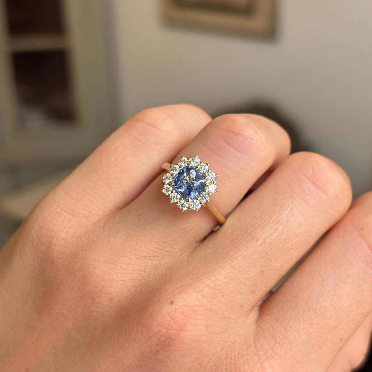 A close-up of a hand wearing a yellow gold ring with a blue sapphire centre stone and a halo of diamonds. Antique-inspired. 