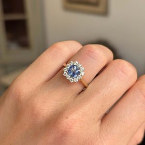 A close-up of a hand wearing a yellow gold ring with a blue sapphire centre stone and a halo of diamonds. Antique-inspired. 