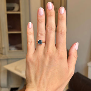 A close-up of a hand wearing an Antique-inspired yellow gold ring with a square-cut sapphire, set against a softly blurred indoor background.
