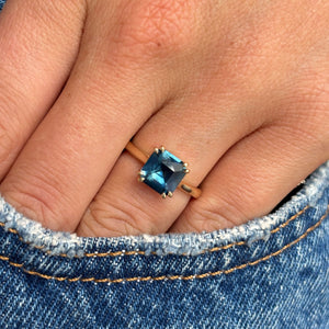 A close-up of a hand wearing an Antique-inspired, teal square-cut sapphire solitaire ring in yellow gold, resting against blue denim fabric.