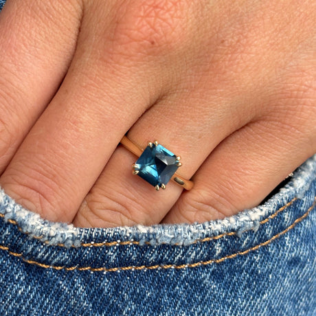 A close-up of a hand wearing an Antique-inspired, teal square-cut sapphire solitaire ring in yellow gold, resting against blue denim fabric.