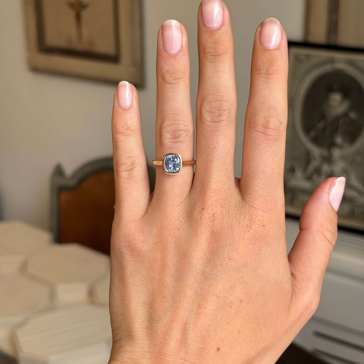 A close-up of a hand wearing an antique-style yellow gold ring with a blue sapphire, set against a softly blurred indoor background.