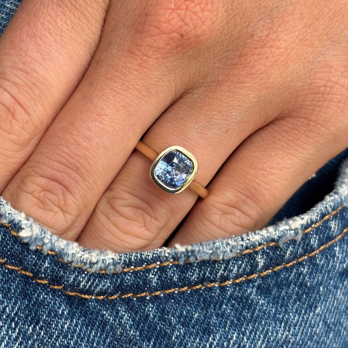 A close-up of a hand wearing a solitaire gold ring set with a square cushion-cut blue sapphire, inspired by Antique designs, resting against blue denim fabric.
