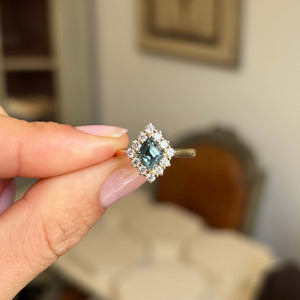 A hand holding an Antique-inspired, Teal kite-cut sapphire and diamond cluster engagement ring in yellow gold with a softly blurred indoor background.