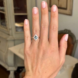 A close-up of a hand wearing an antique-inspired diamond ring with a kite-cut blue sapphire and a halo of smaller diamonds on a gold band, set against a softly blurred indoor background.