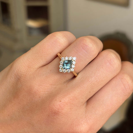 A close-up of a hand wearing a kite cluster antique inspired gold ring featuring a teal sapphire surrounded by diamonds stones, with a softly blurred indoor background.