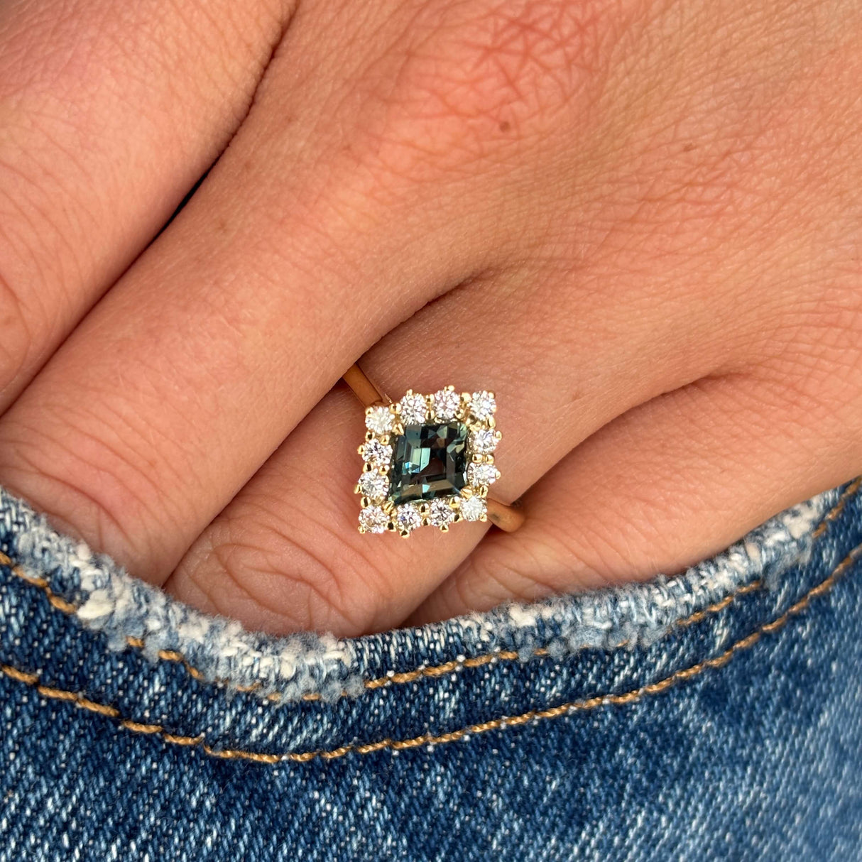 A close-up of a hand wearing an Antique-inspired, Teal kite-cut sapphire and diamond cluster engagement ring with blue denim visible at the bottom.