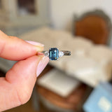 Hand holding a Vintage-inspired, Teal sapphire and diamond three-stone engagement ring in platinum against a blurred indoor background