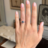 Hand wearing a platinum Vintage-inspired, Teal sapphire and diamond three-stone engagement ring against a blurred indoor background
