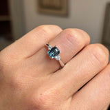 Vintage-inspired, Teal sapphire and diamond three-stone engagement ring in platinum on a person's finger against a neutral background