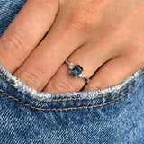 Hand wearing a platinum Vintage-inspired, Teal sapphire and diamond three-stone engagement ring against a denim background