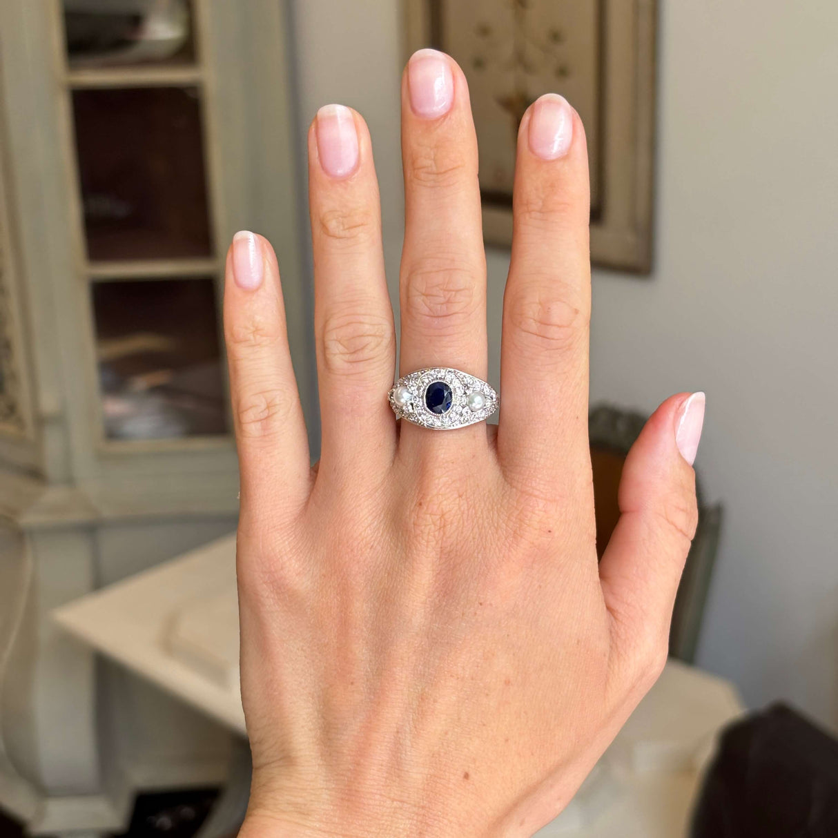 Hand wearing an Antique, Belle Époque sapphire, pearl and diamond cluster cocktail ring in white gold against a blurred indoor background