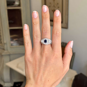 Hand wearing an Antique, Belle Époque sapphire, pearl and diamond cluster cocktail ring in white gold against a blurred indoor background