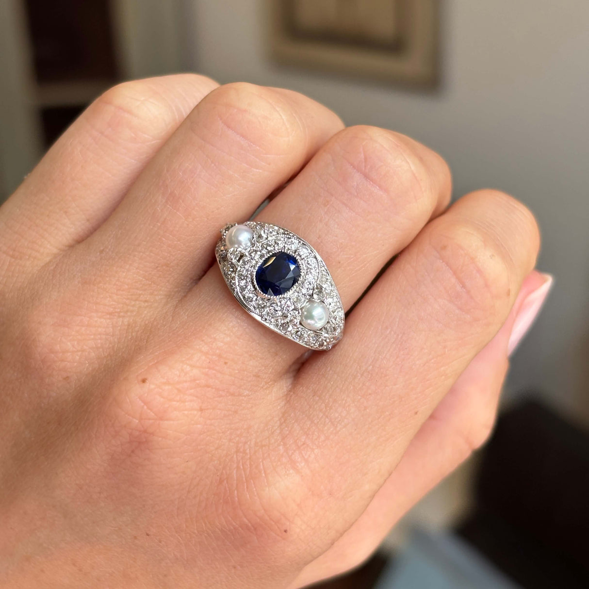 An Antique, Belle Époque dark blue sapphire, pearl and diamond cluster cocktail ring in white gold worn on a person's finger.