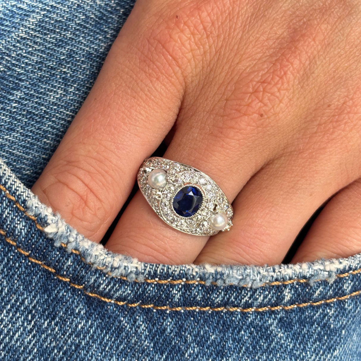 Close-up of a hand wearing a white gold Antique, Belle Époque sapphire, pearl and diamond cluster cocktail ring against a denim background.