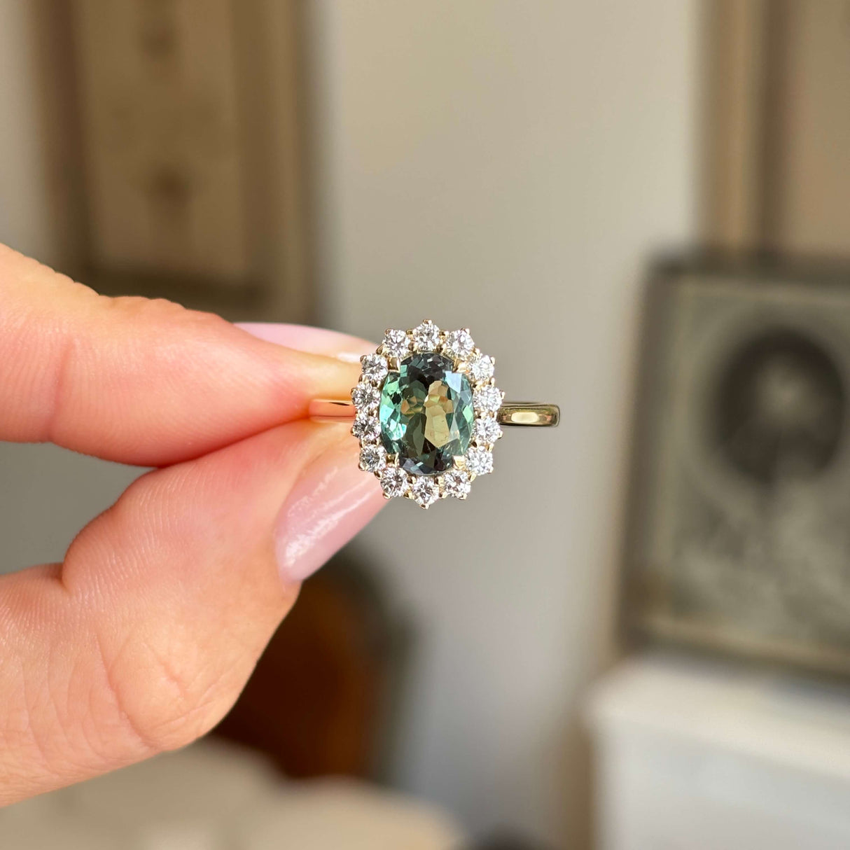 Hand holding an Antique-inspired, gold ring with a central colour-change alexandrite surrounded by a halo of round-brilliant diamonds. 