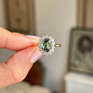 Hand holding an Antique-inspired, gold ring with a central colour-change alexandrite surrounded by a halo of round-brilliant diamonds. 