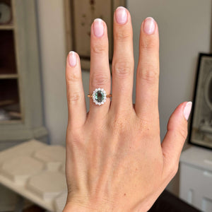 Hand wearing a ring with an Antique-inspired, alexandrite and diamond cluster engagement ring against a neutral background