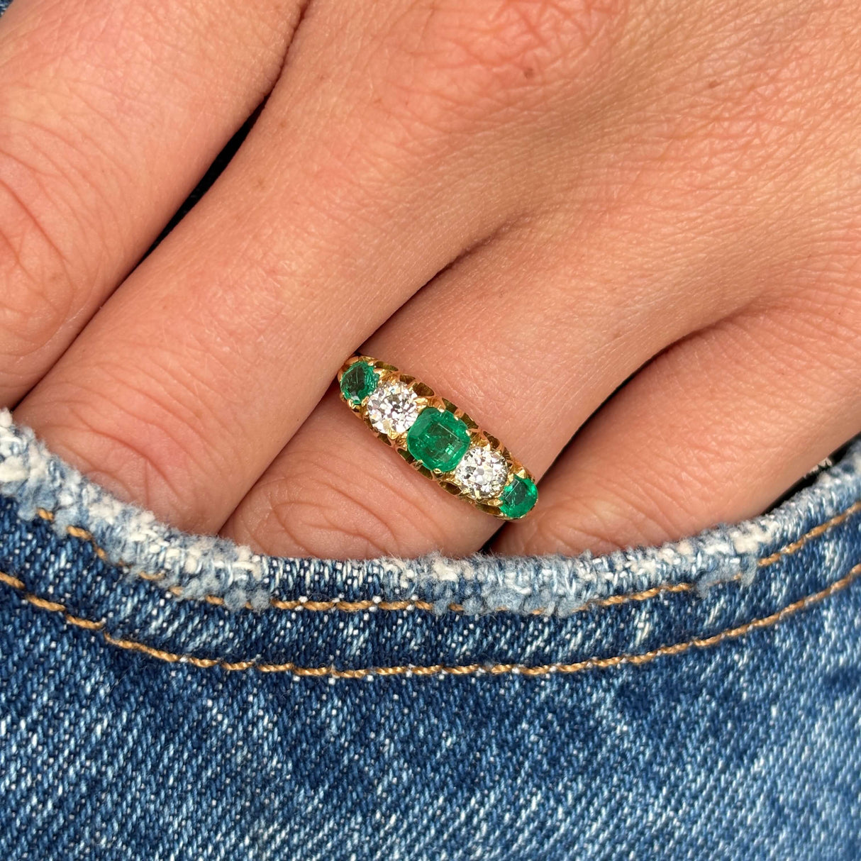 Close up of an Antique, Victorian gold five-stone ring with three old-mine emeralds and two old-mine cut diamonds worn on a finger against a denim background