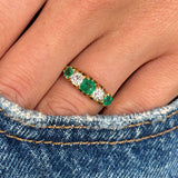 Close up of an Antique, Victorian gold five-stone ring with three old-mine emeralds and two old-mine cut diamonds worn on a finger against a denim background