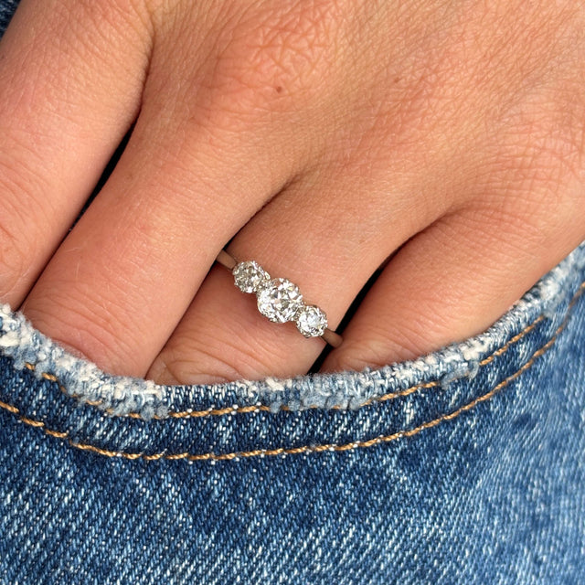 A three-stone vintage white gold engagement ring worn on a hand against a denim background, showcasing the three old-cut diamonds. 