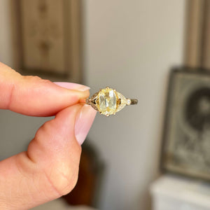 Vintage-inspired, yellow cushion-cut sapphire and diamond engagement ring in yellow gold, held between two fingers against a blurred interior backdrop