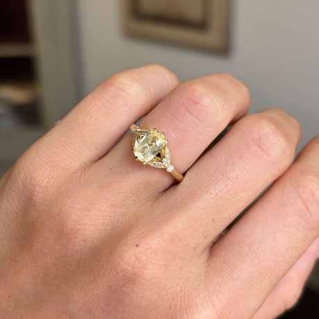 Close up of a yellow cushion-cut sapphire set with diamonds in a yellow gold ring, vintage inspired and worn on a hand.