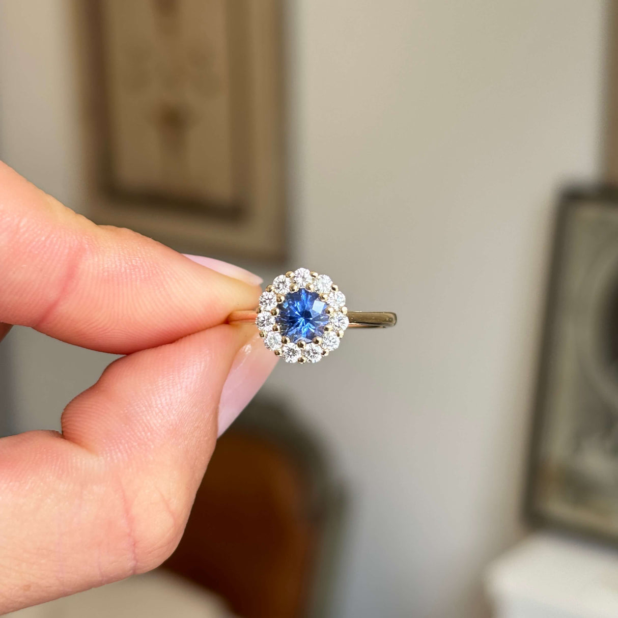 Antique-inspired, blue sapphire and diamond cluster engagement ring in yellow gold, held by a hand against a neutral background