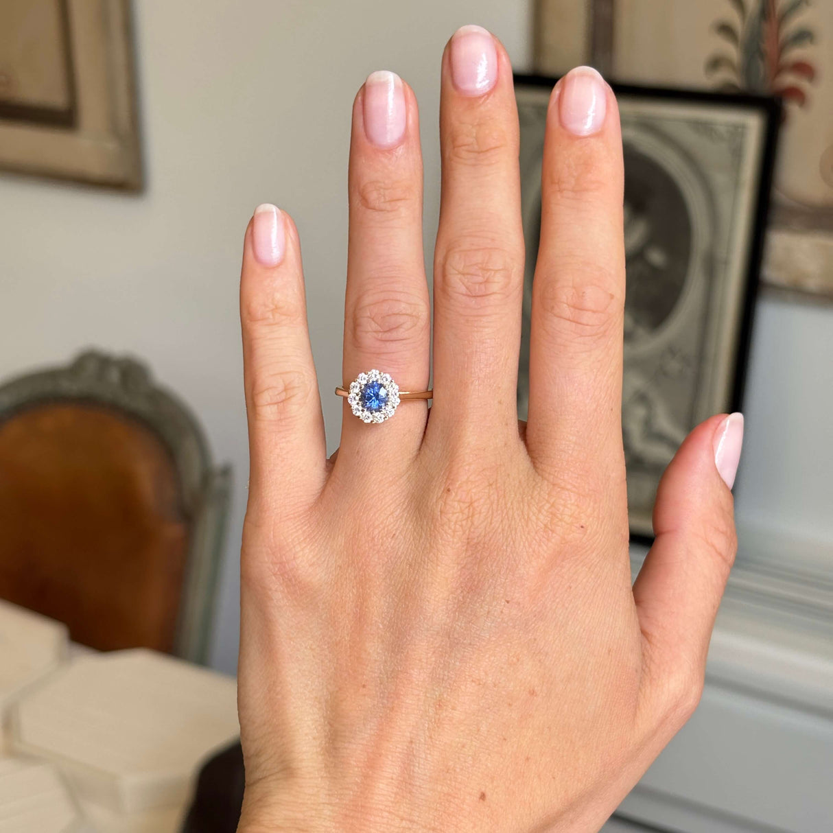 Hand wearing a gold Antique-inspired, blue sapphire and diamond cluster engagement ring in a decorative indoor setting