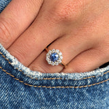 Close-up of a hand wearing an Antique-inspired, electric blue sapphire and diamond cluster engagement ring in yellow gold on denim fabric.