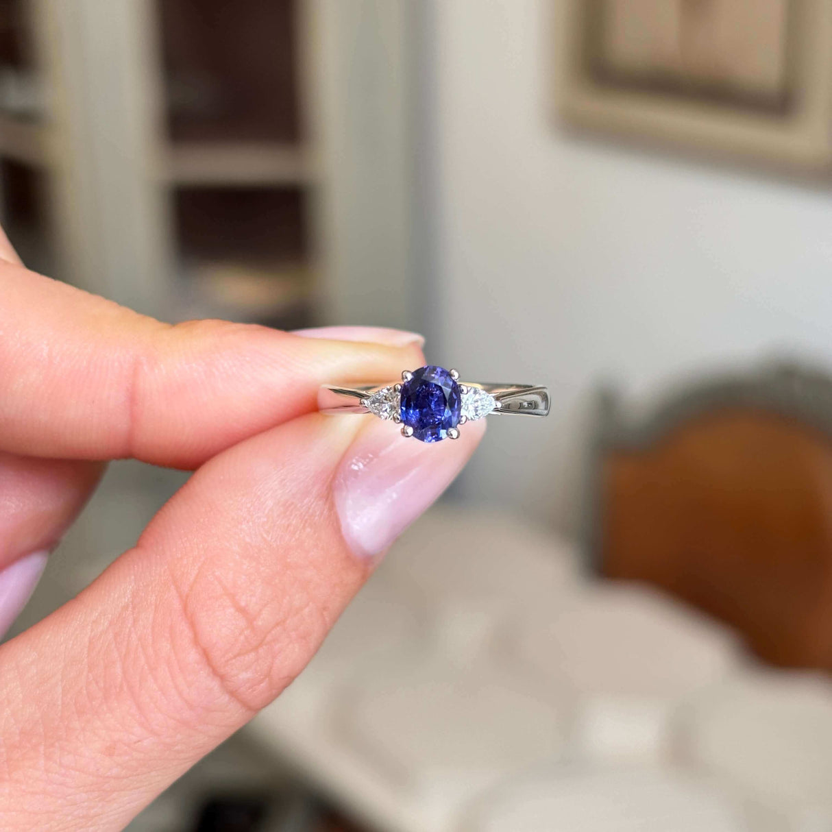 Hand holding a vintage-inspired, deep indigo sapphire & diamond three-stone engagement ring in platinum against a blurred indoor background