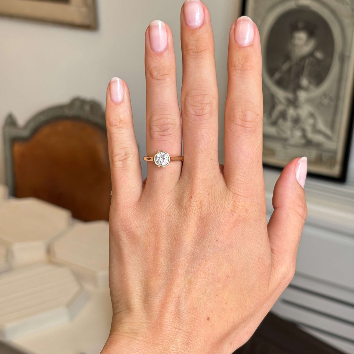 A close-up of a hand wearing a delicate yellow-gold solitaire engagement ring with a round diamond in antique-inspired bezel setting against a softly blurred indoor background.