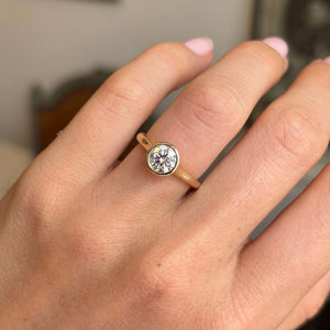 A close up of a 'salt and pepper' diamond in an antique-inspired solitaire engagement ring, worn on a hand showcasing the yellow gold rub-over setting and band, against a softly blurred background. 