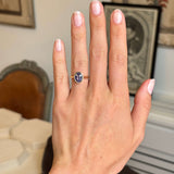 A hand wearing an Antique-inspired, purple oval sapphire solitaire engagement ring, set against a softly blurred indoor background.