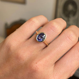 A gold antique-inspired solitaire engagement ring set with a purple sapphire in a bezel setting worn on a person's finger, shown in a close-up shot.