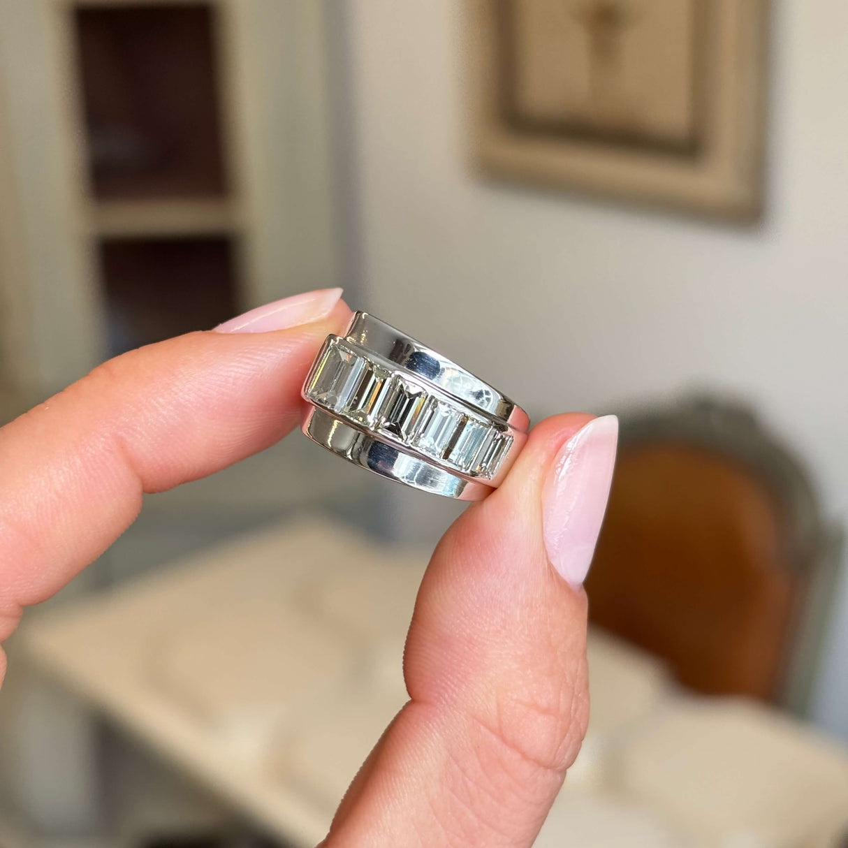 Vintage, 1940s channel-set baguette-cut diamond platinum ring held between fingers with a blurred indoor background