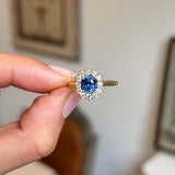 A hand holding an Antique-inspired, blue sapphire and diamond cluster engagement ring in yellow gold, set against a softly blurred indoor background.