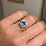 A close-up of a hand wearing an antique-inspired yellow gold ring with a central blue sapphire surrounded by a halo of diamonds. 