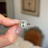 Antique inspired, yellow sapphire and diamond cluster engagement ring in platinum held by a hand against a blurred indoor background
