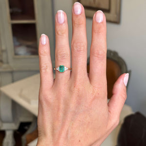 Hand wearing an Antique-inspired, Emerald and diamond three-stone engagement ring against a blurred indoor background