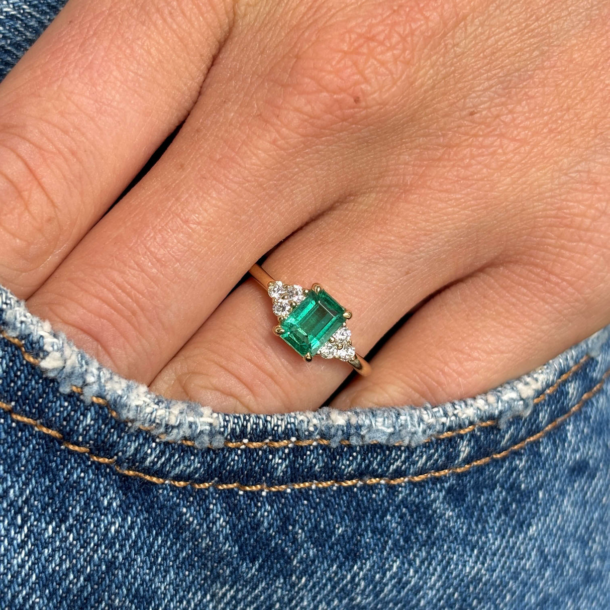 Antique-inspired, Emerald and diamond three-stone yellow gold engagement ring on a finger against a denim background