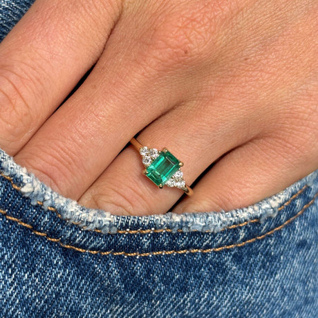 Antique-inspired, Emerald and diamond three-stone yellow gold engagement ring on a finger against a denim background
