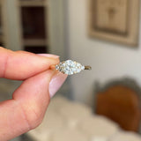 Antique-inspired, cloudy diamond cluster trefoil ring held between fingers with a blurred indoor background