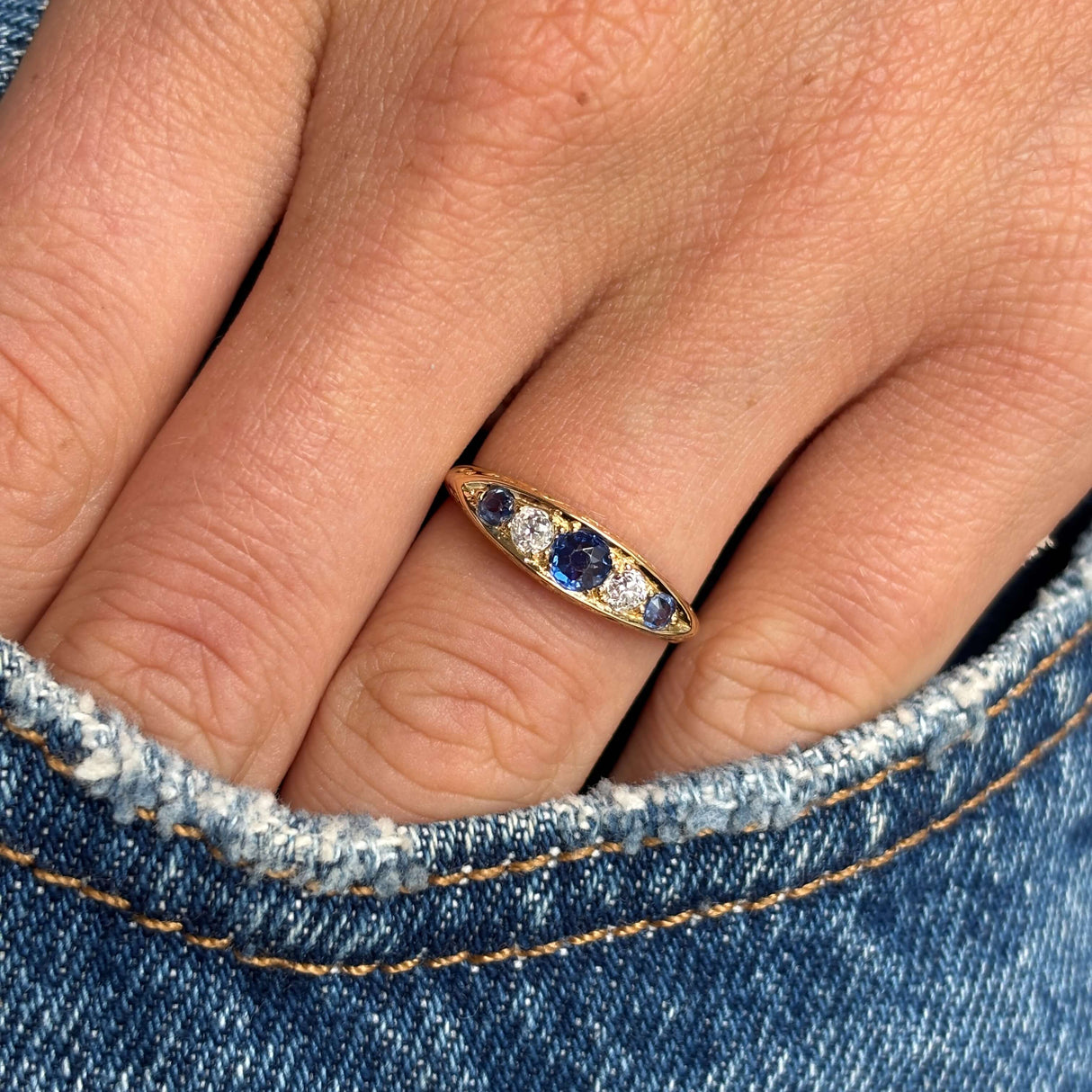 Close-up of a hand wearing an Antique, Victorian sapphire & diamond five-stone engagement ring in gold, against a denim background.