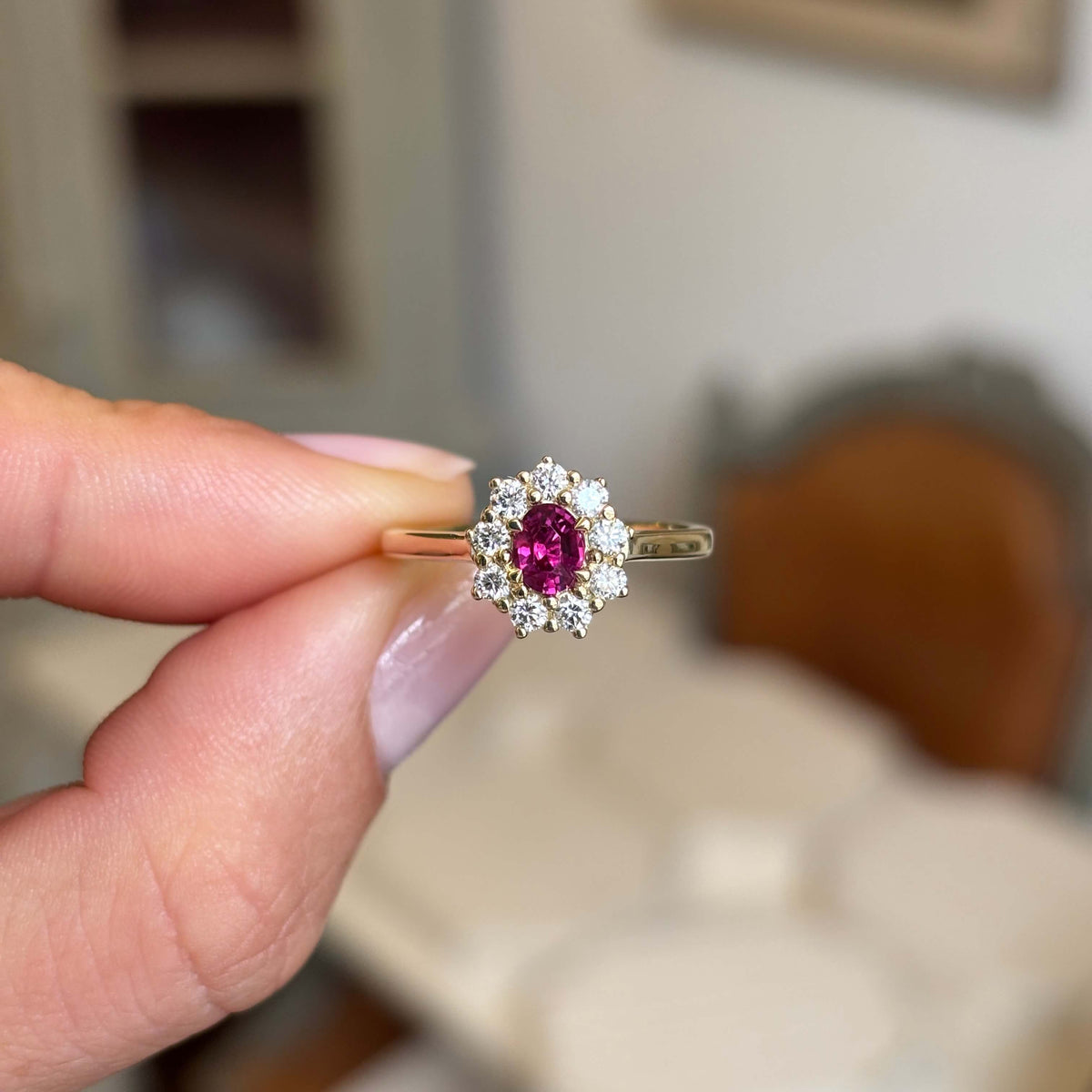 A hand holding a yellow gold antique-inspired engagement ring with a central oval pink ruby surrounded by diamonds, set against a blurred indoor background.