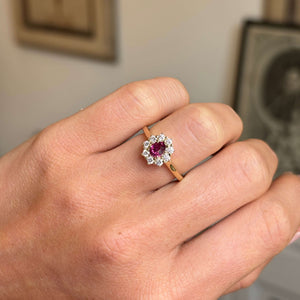 A close-up of a hand wearing a yellow gold antique-inspired gold engagement ring with a central oval pinkish-red ruby surrounded by a halo of diamonds. 