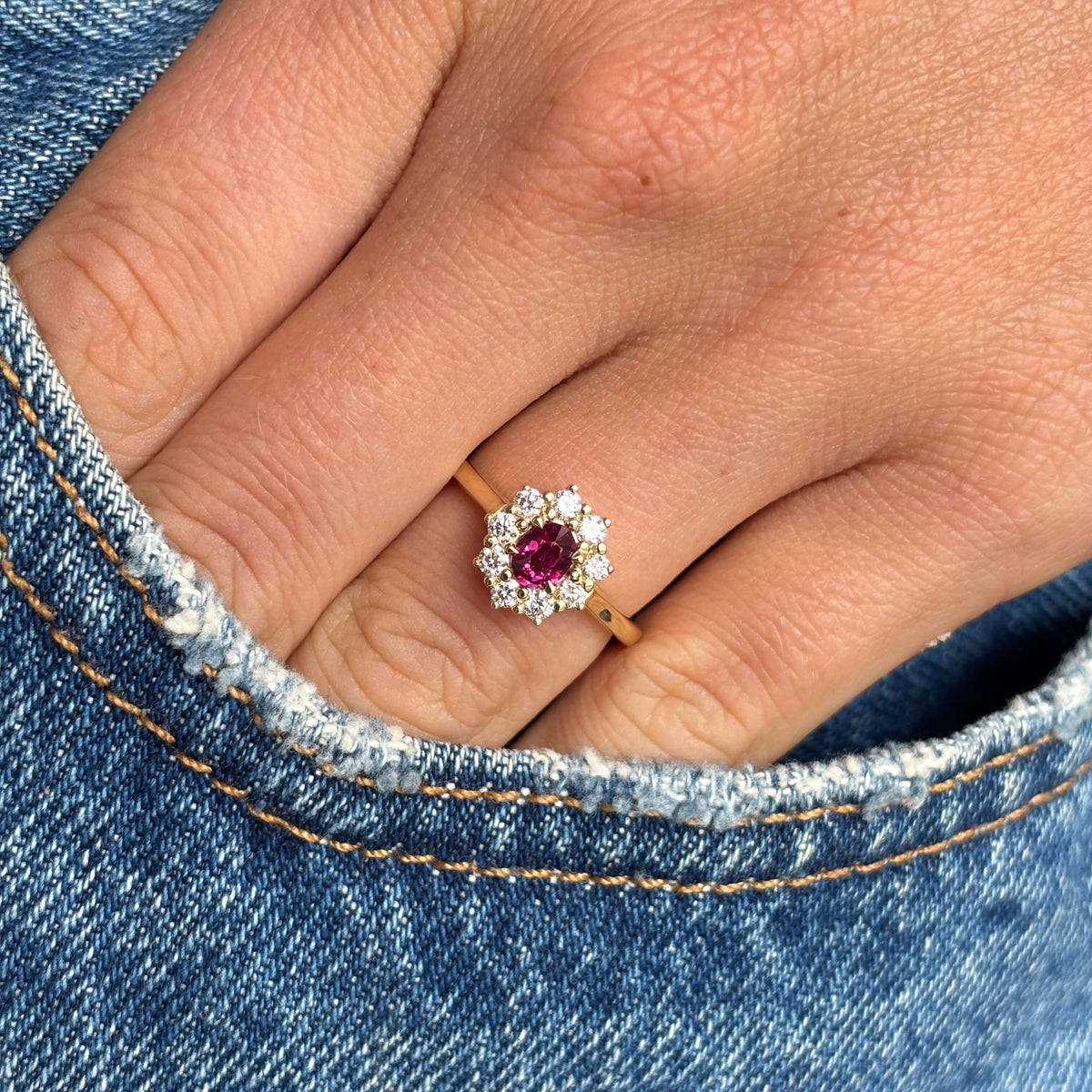 A close-up of a hand wearing a yellow gold ring with a central round pink ruby surrounded by a halo of diamonds, set against blue denim fabric.